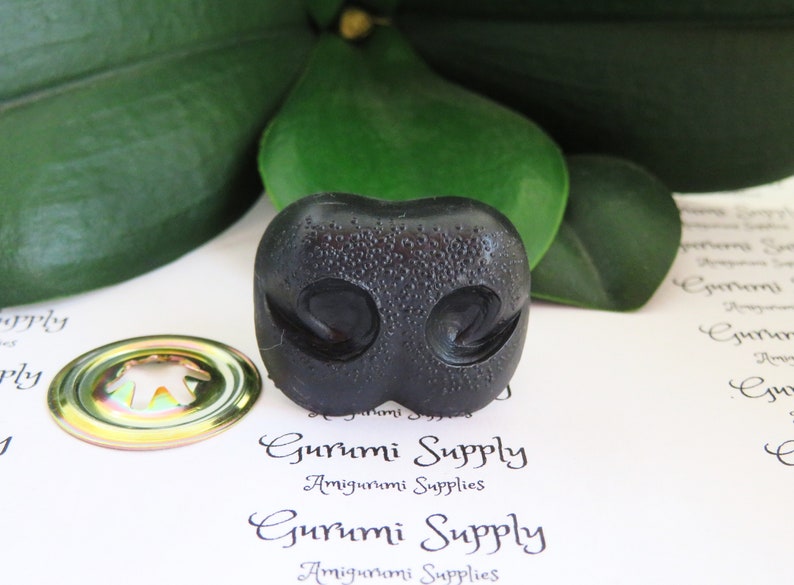 May include: Black plastic amigurumi nose with a textured surface. The nose has two nostrils and a small indentation in the center.