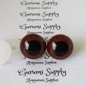 Puede incluir: Dos ojos de seguridad marrones con pupilas negras y arandelas blancas. Los ojos tienen un di&aacute;metro de 12 mm. Las arandelas tienen un di&aacute;metro de 18 mm. Los ojos est&aacute;n sobre un fondo blanco con el texto "Gurumi Supply Amigurumi Supplies" impreso en &eacute;l.