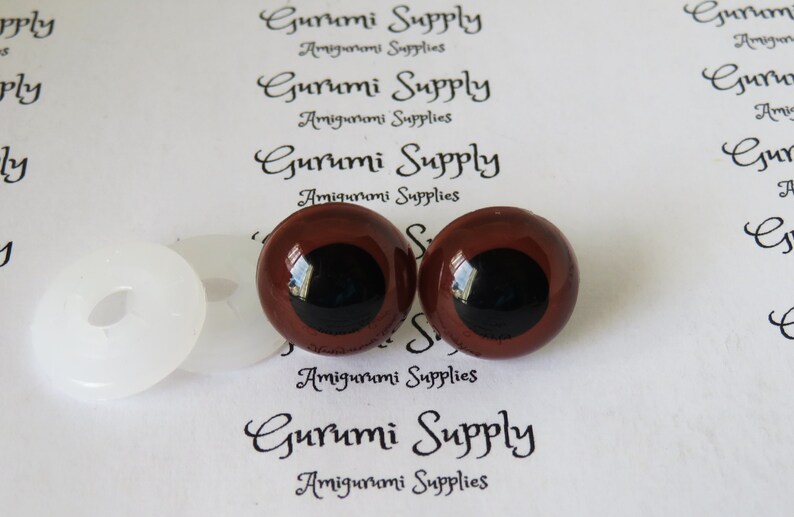 Puede incluir: Dos ojos de seguridad marrones con pupilas negras y arandelas blancas. Los ojos tienen un di&aacute;metro de 12 mm. Las arandelas tienen un di&aacute;metro de 18 mm. Los ojos est&aacute;n sobre un fondo blanco con el texto "Gurumi Supply Amigurumi Supplies" impreso en &eacute;l.