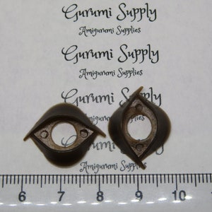 May include: Two dark brown, eye-shaped amigurumi safety eyes. Each eye has a circular center and small holes for attachment. The text "Gurumi Supply Amigurumi Supplies" is visible in the background.