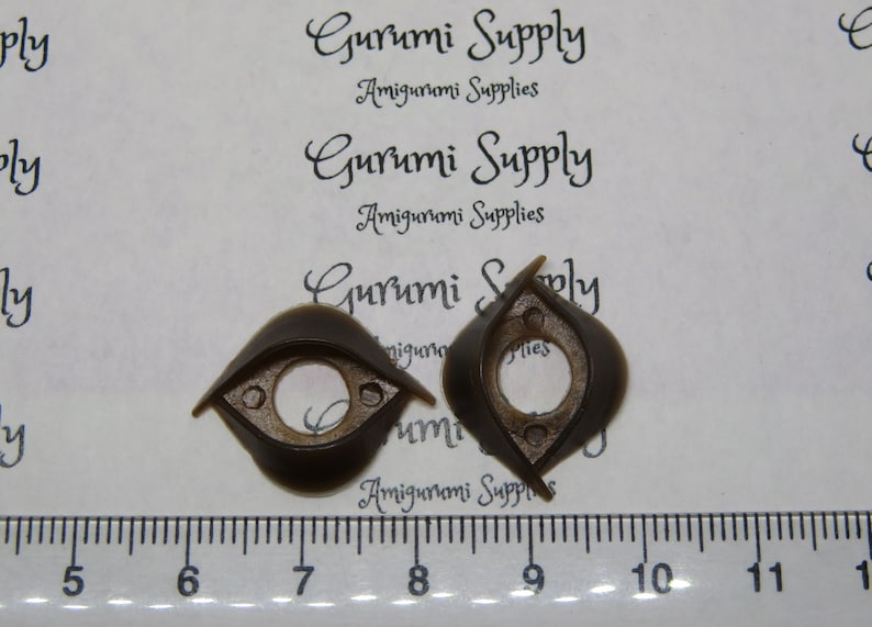 May include: Two dark brown, eye-shaped amigurumi safety eyes. Each eye has a circular center and small holes for attachment. The text "Gurumi Supply Amigurumi Supplies" is visible in the background.