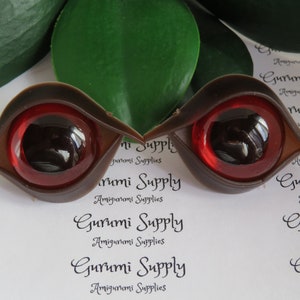 Puede incluir: Dos ojos de mu&ntilde;eca de pl&aacute;stico marr&oacute;n con iris rojos y pupilas negras. Los ojos tienen forma de l&aacute;grima y un acabado brillante. El texto "Gurumi Supply" est&aacute; impreso en el fondo.