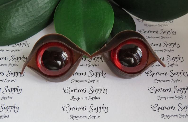 Puede incluir: Dos ojos de mu&ntilde;eca de pl&aacute;stico marr&oacute;n con iris rojos y pupilas negras. Los ojos tienen forma de l&aacute;grima y un acabado brillante. El texto "Gurumi Supply" est&aacute; impreso en el fondo.
