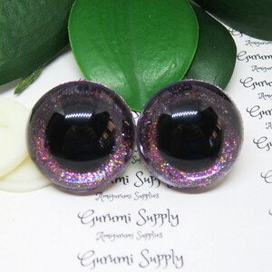 May include: Two purple and black glitter safety eyes with a black pupil. The eyes are on a white background with the text "Gurumi Supply" printed on it.