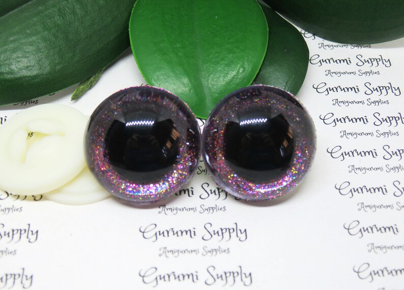 May include: Two purple and black glitter safety eyes with a black pupil. The eyes are on a white background with the text "Gurumi Supply" printed on it.