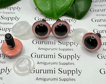 Puede incluir: Un conjunto de ojos de seguridad para amigurumi, con pupilas negras e iris marrón-rosado. También se incluyen arandelas de plástico transparente y postes atornillables. El texto "Gurumi Supply" es visible.