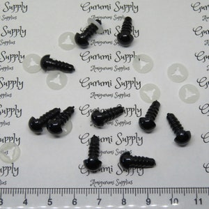 May include: A close-up of ten black plastic safety eyes with a screw back. The eyes are 12mm in diameter and are arranged in a scattered pattern on a white surface. A ruler is visible in the bottom of the image with markings from 3 to 11.