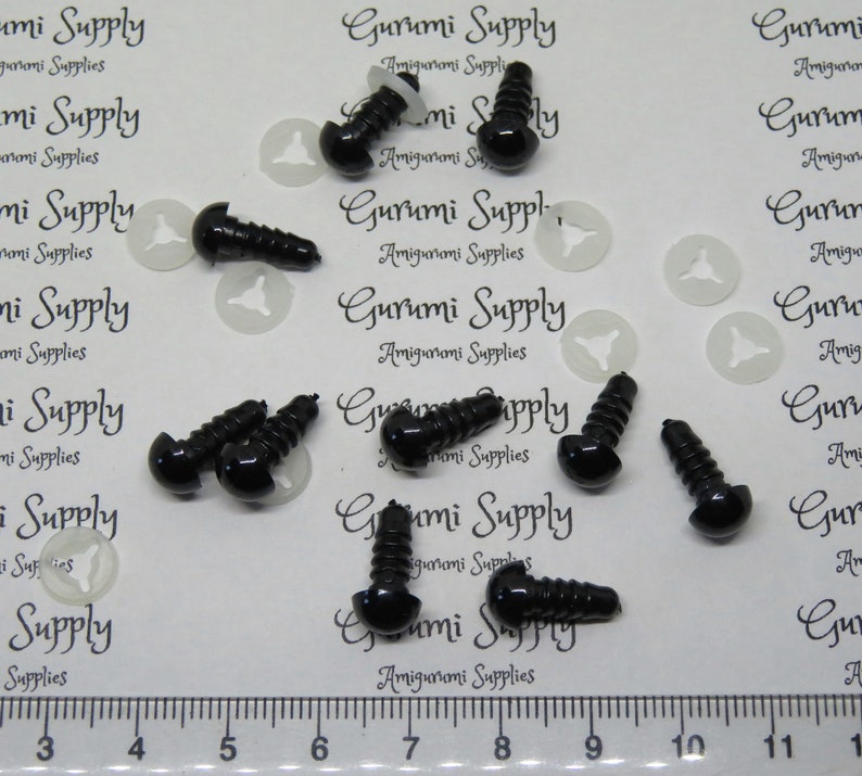 May include: A close-up of ten black plastic safety eyes with a screw back. The eyes are 12mm in diameter and are arranged in a scattered pattern on a white surface. A ruler is visible in the bottom of the image with markings from 3 to 11.