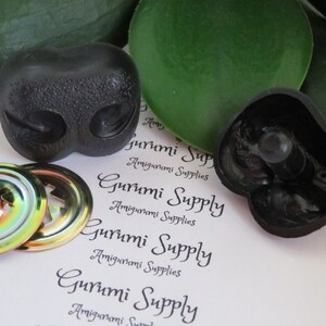 May include: Black plastic amigurumi animal nose with two holes for eyes. Two iridescent metal safety eyes with a black plastic backing. The image also includes the text "Gurumi Supply Amigurumi Supplies".