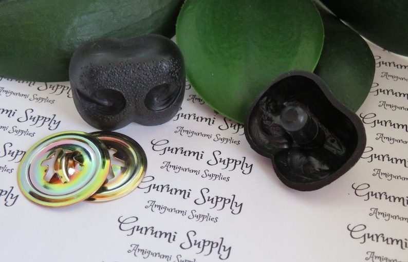 May include: Black plastic amigurumi animal nose with two holes for eyes. Two iridescent metal safety eyes with a black plastic backing. The image also includes the text "Gurumi Supply Amigurumi Supplies".