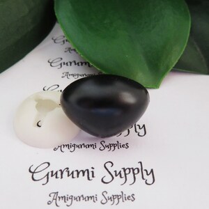 May include: Two plastic safety eyes for amigurumi projects. One eye is black and the other is white. The eyes are on a white surface with the text "Gurumi Supply" printed on it.