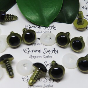 Puede incluir: Primer plano de una colecci&oacute;n de ojos de seguridad de pl&aacute;stico verde y negro con tornillos de metal dorado. Los ojos est&aacute;n dispuestos sobre una superficie blanca con el texto "Gurumi Supply" impreso en ella.