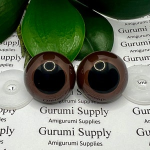 Puede incluir: Dos ojos de seguridad marrones con pupilas negras y un anillo dorado alrededor de la pupila. Los ojos están sobre una superficie blanca con el texto "Gurumi Supply" impreso en ella.