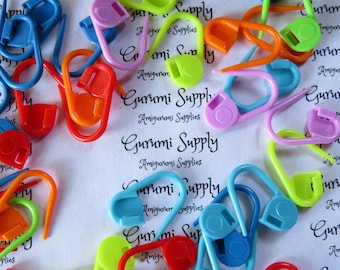 May include: Assortment of colorful plastic stitch markers in various shapes, including circles and hooks. The markers are used for counting rows or stitches in knitting and crocheting projects. The text "Gurumi Supply Amigurumi Supplies" is printed on the white background.
