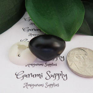 May include: A close-up of a black plastic safety eye for amigurumi projects. The eye is next to a white plastic safety eye and a US quarter for size comparison. The text "Gurumi Supply" is visible in the background.