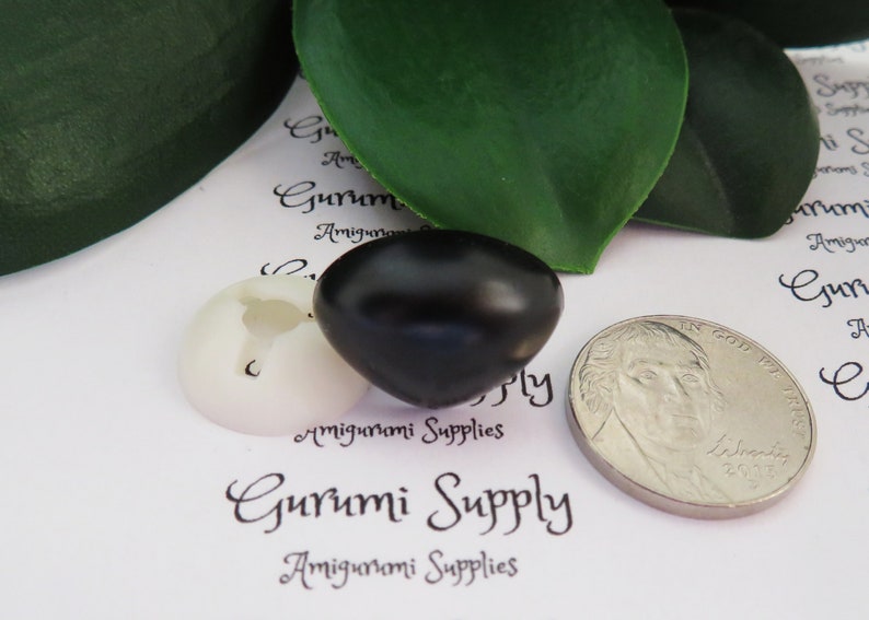 May include: A close-up of a black plastic safety eye for amigurumi projects. The eye is next to a white plastic safety eye and a US quarter for size comparison. The text "Gurumi Supply" is visible in the background.
