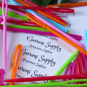 May include: A close-up of a variety of colorful plastic yarn needles, also known as tapestry needles, with the text "Gurumi Supply Amigurumi Supplies" printed on a white background.