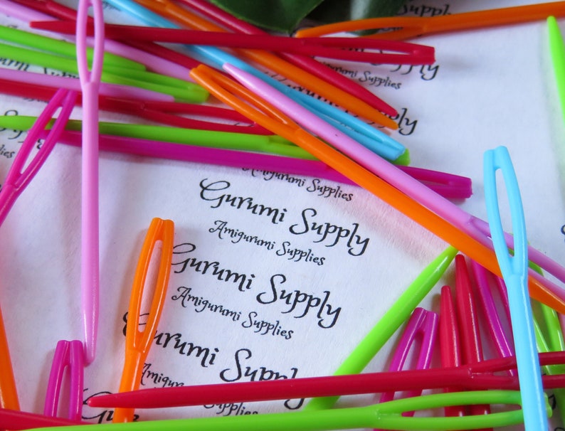 May include: A close-up of a variety of colorful plastic yarn needles, also known as tapestry needles, with the text "Gurumi Supply Amigurumi Supplies" printed on a white background.
