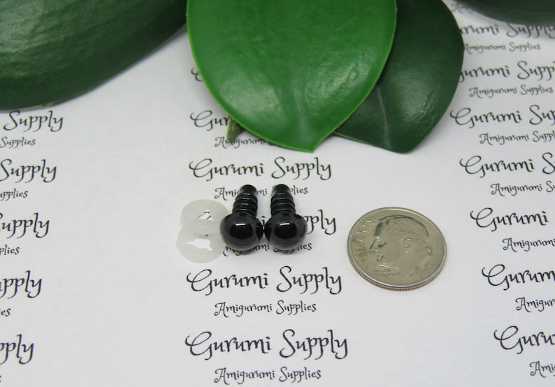May include: Two black plastic safety eyes with a white plastic washer, a silver dime for size comparison, and the text "Gurumi Supply Amigurumi Supplies" printed on a white background.