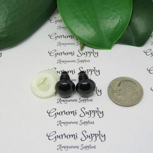 K&ouml;nnte beinhalten: Zwei schwarze Sicherheitsaugen aus Kunststoff mit einer wei&szlig;en Kunststoffscheibe, neben einer US-Dime-M&uuml;nze zum Gr&ouml;&szlig;envergleich.  Gurumi Supply Amigurumi Supplies ist auf dem wei&szlig;en Hintergrund gedruckt.