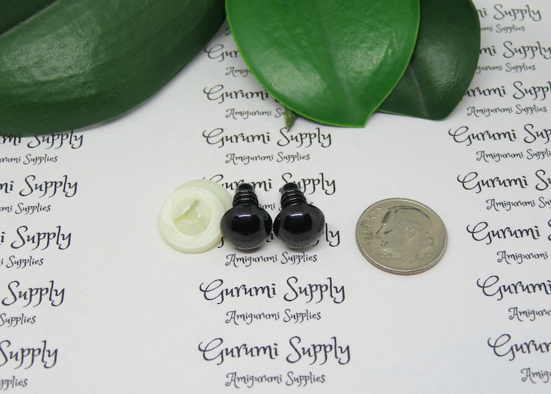 K&ouml;nnte beinhalten: Zwei schwarze Sicherheitsaugen aus Kunststoff mit einer wei&szlig;en Kunststoffscheibe, neben einer US-Dime-M&uuml;nze zum Gr&ouml;&szlig;envergleich.  Gurumi Supply Amigurumi Supplies ist auf dem wei&szlig;en Hintergrund gedruckt.