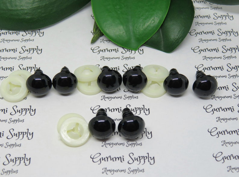 May include: A close-up of six black plastic safety eyes with white plastic washers. The eyes are for use in amigurumi and other crafts.