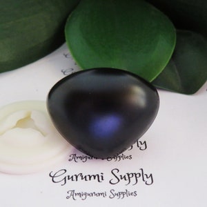 May include: A close-up of a black, heart-shaped safety eye for amigurumi projects. The eye is sitting on a white surface with the text "Gurumi Supply" printed on it.