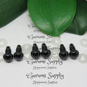 May include: A close-up of ten black plastic safety eyes with a screw back, used for amigurumi projects. The eyes are arranged in two rows of five, with white plastic washers in between.