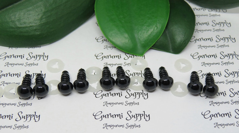 May include: A close-up of ten black plastic safety eyes with a screw back, used for amigurumi projects. The eyes are arranged in two rows of five, with white plastic washers in between.