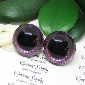 May include: Two black and purple glitter safety eyes with a black pupil. The eyes are round and have a shiny finish.