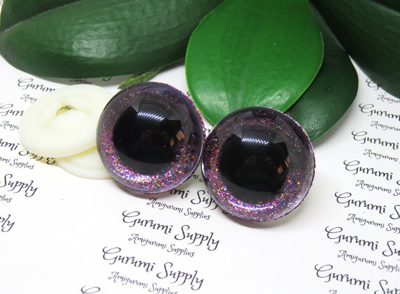 May include: Two black and purple glitter safety eyes with a black pupil. The eyes are round and have a shiny finish.