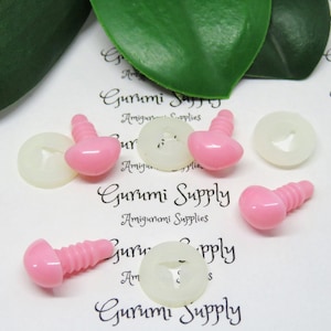 May include: Pink plastic safety eyes with black screw backs and white plastic washers. The eyes are on a white surface with the text "Gurumi Supply Amigurumi Supplies" printed on it.