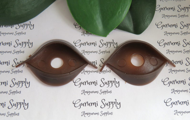 Puede incluir: Dos ojos de seguridad de pl&aacute;stico marr&oacute;n para amigurumi u otras manualidades. Cada ojo tiene una forma redondeada con un agujero central y una peque&ntilde;a pesta&ntilde;a. El texto "Gurumi Supply" es visible en el fondo.