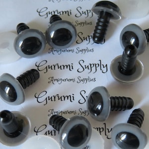 Puede incluir: Primer plano de un grupo de ojos de seguridad de plástico gris con pupilas negras y tornillos negros de plástico. Los ojos están esparcidos sobre una superficie blanca con el texto "Gurumi Supply" impreso en ella.
