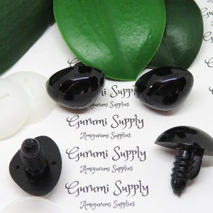 May include: A close-up of black plastic safety eyes for amigurumi projects. The eyes are oval shaped and have a screw back. The eyes are on a white background with the text "Gurumi Supply Amigurumi Supplies" printed on it.