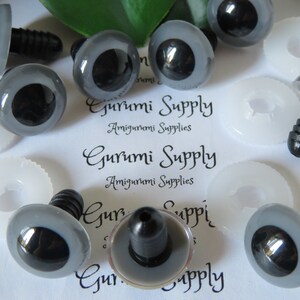 May include: A close-up of a set of gray plastic safety eyes with black pupils. The eyes are on a white surface with the text "Gurumi Supply Amigurumi Supplies" printed on it.