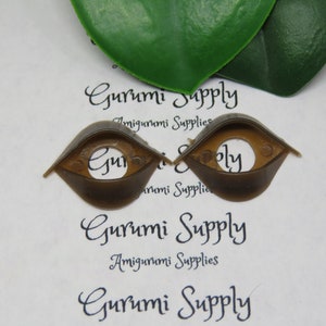 May include: Two brown plastic amigurumi safety eyes with a circular center hole. The eyes are in a teardrop shape. The text "Gurumi Supply" and "Amigurumi Supplies" is printed on the background.
