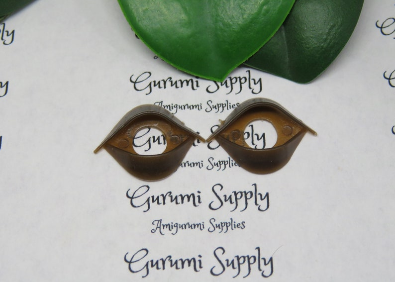 May include: Two brown plastic amigurumi safety eyes with a circular center hole. The eyes are in a teardrop shape. The text "Gurumi Supply" and "Amigurumi Supplies" is printed on the background.
