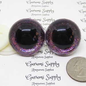 May include: Two black and purple glitter safety eyes with a black pupil. The eyes are approximately 1 inch in diameter. The eyes are on a white background with the text "Gurumi Supply" printed on it.