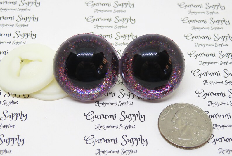 May include: Two black and purple glitter safety eyes with a black pupil. The eyes are approximately 1 inch in diameter. The eyes are on a white background with the text "Gurumi Supply" printed on it.