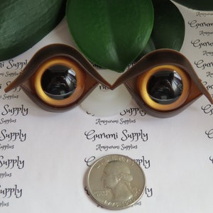 Puede incluir: Dos ojos de mu&ntilde;eca de pl&aacute;stico marr&oacute;n con iris dorado y pupila negra. Los ojos tienen forma de almendra estilizada. Se incluye una moneda como referencia de tama&ntilde;o. El texto "Gurumi Supply" est&aacute; impreso.