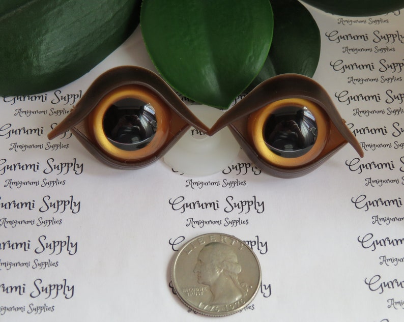 Puede incluir: Dos ojos de mu&ntilde;eca de pl&aacute;stico marr&oacute;n con iris dorado y pupila negra. Los ojos tienen forma de almendra estilizada. Se incluye una moneda como referencia de tama&ntilde;o. El texto "Gurumi Supply" est&aacute; impreso.