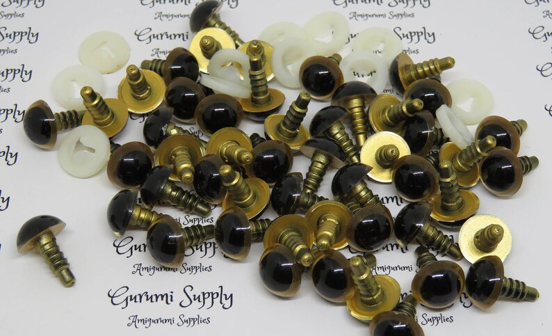 Puede incluir: Primer plano de un mont&oacute;n de ojos de seguridad de pl&aacute;stico negro y marr&oacute;n con tornillos de metal dorado. Los ojos se utilizan para amigurumi y otros proyectos de artesan&iacute;a.