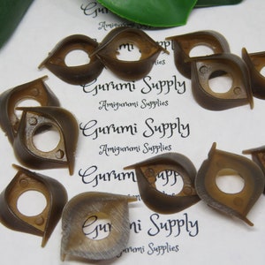 May include: A collection of brown plastic doll eyes with a unique, stylized shape. The eyes have a central hole and are designed for amigurumi projects. The text "Gurumi Supply Amigurumi Supplies" is visible.