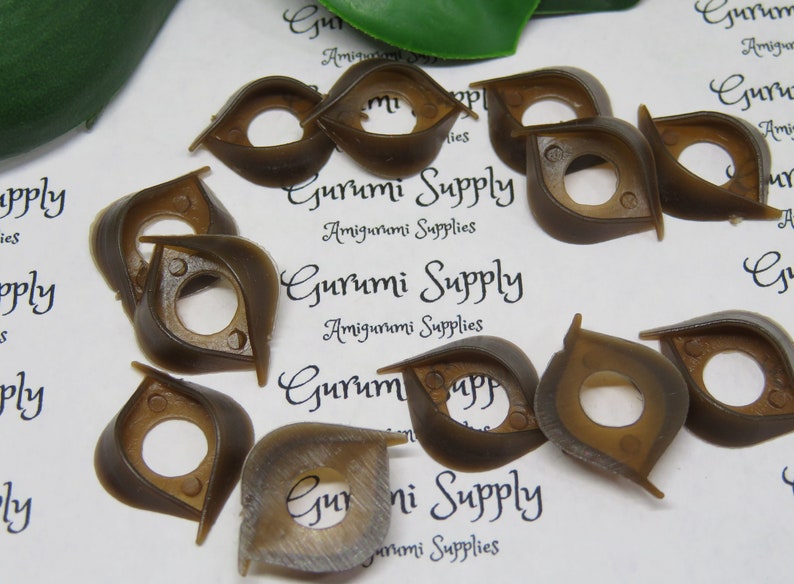 May include: A collection of brown plastic doll eyes with a unique, stylized shape. The eyes have a central hole and are designed for amigurumi projects. The text "Gurumi Supply Amigurumi Supplies" is visible.