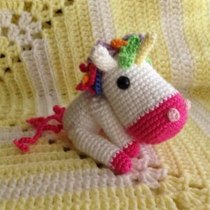 May include: A white crocheted unicorn with a pink tail and a rainbow mane. The unicorn is sitting on a yellow and white crocheted blanket.