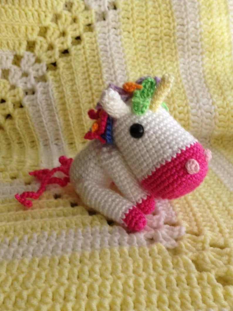 May include: A white crocheted unicorn with a pink tail and a rainbow mane. The unicorn is sitting on a yellow and white crocheted blanket.
