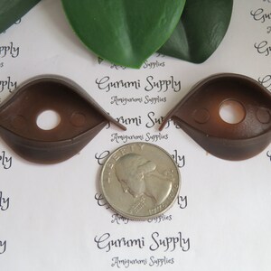 Puede incluir: Dos ojos de seguridad de pl&aacute;stico marr&oacute;n para proyectos de amigurumi, cada uno con un agujero central y dos peque&ntilde;os agujeros laterales. Se coloca una moneda entre los ojos para comparar el tama&ntilde;o. El texto "Gurumi Supply Amigurumi Supplies" est&aacute; impreso en el fondo.