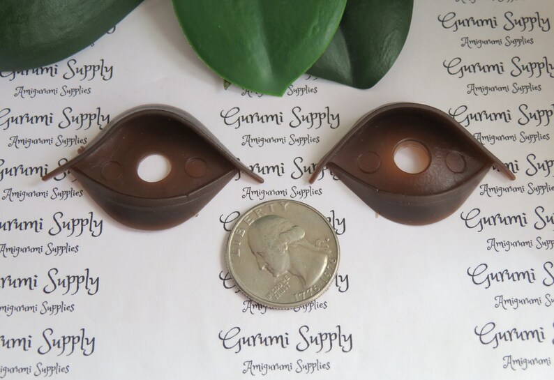 Puede incluir: Dos ojos de seguridad de pl&aacute;stico marr&oacute;n para proyectos de amigurumi, cada uno con un agujero central y dos peque&ntilde;os agujeros laterales. Se coloca una moneda entre los ojos para comparar el tama&ntilde;o. El texto "Gurumi Supply Amigurumi Supplies" est&aacute; impreso en el fondo.