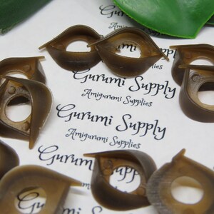 May include: A collection of brown plastic safety eyes for amigurumi projects. The eyes have a unique shape with a central hole and a curved, wing-like design. The text "Gurumi Supply" and "Amigurumi Supplies" is visible.